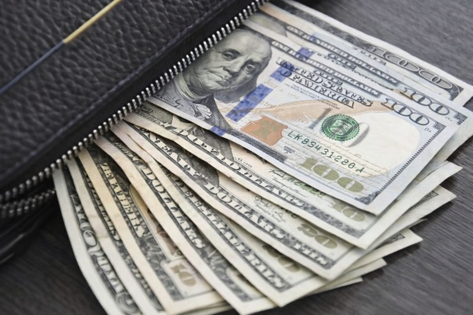 A black leather wallet partially unzipped, with neatly fanned U.S. dollar bills inside, placed on a dark wooden surface.